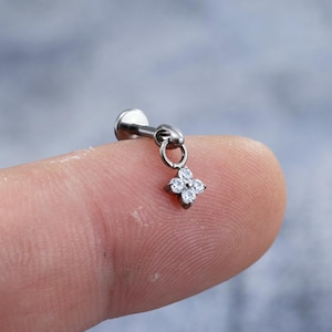 Pode incluir: Um piercing labret prateado com um pendente de flor de quatro pétalas. A flor é adornada com quatro pedras preciosas redondas e transparentes. A joia é mostrada num dedo, destacando o seu tamanho pequeno e os seus detalhes intrincados.