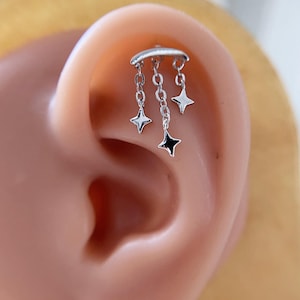 Può includere: Un piercing all'orecchio argentato con una barra curva e tre catenine pendenti, ciascuna con un ciondolo a forma di stella. Le stelle hanno una finitura lucida, una con il centro nero. Il gioiello è mostrato su un orecchio.