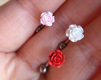May include: Three rose-shaped body jewelry pieces. The roses are made of a white material and are in pink, white, and red.