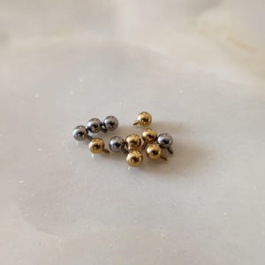 May include: A selection of small, metallic body jewellery. The image shows several silver and gold-coloured ball-shaped studs with screw-on posts. The jewellery is scattered on a light-coloured surface.