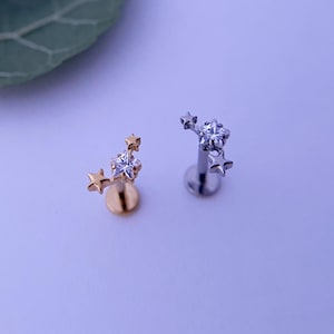 Puede incluir: Dos pendientes de labio o de oreja con forma de estrella. Un pendiente es de color dorado con una piedra central clara y brillante y tres estrellas más pequeñas. El otro pendiente es de color plateado con una piedra central clara y brillante y tres estrellas más pequeñas. Los pendientes están sobre una superficie de color púrpura claro.