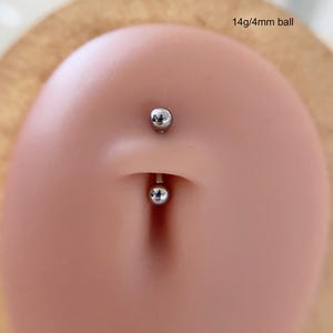 May include: A silver belly button ring with two small, round balls. The top ball is above the navel, and the bottom ball is below. The text "14g/4mm ball" is visible.