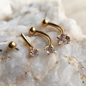 Puede incluir: Tres piezas de joyer&iacute;a corporal de barra curva de color dorado con acentos de cristal transparente. La joyer&iacute;a se muestra sobre una superficie de roca blanca.