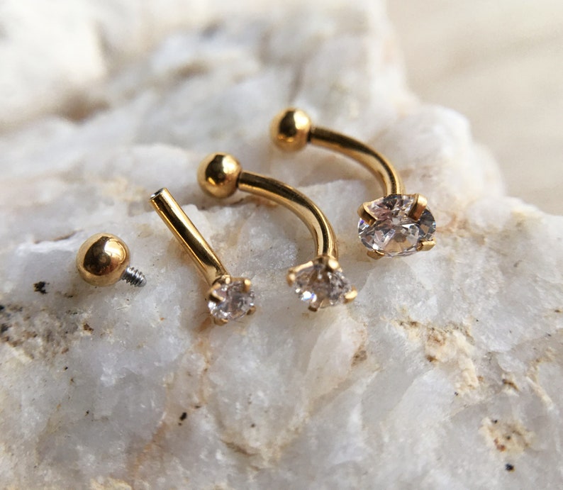 Puede incluir: Tres piezas de joyer&iacute;a corporal de barra curva de color dorado con acentos de cristal transparente. La joyer&iacute;a se muestra sobre una superficie de roca blanca.