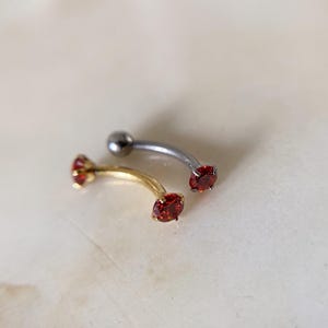 Op de afbeelding: Twee gebogen barbell lichaamsieraden. Het ene sieraad is goudkleurig met rode ronde edelstenen aan beide uiteinden. Het tweede sieraad is zilverkleurig met een gladde metalen bal aan één uiteinde en een rode ronde edelsteen aan het andere.