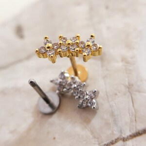 May include: Two gold and silver body jewelry pieces with clear crystal accents. The gold piece is a flower-shaped stud with a post and a clear crystal center. The silver piece is a flower-shaped stud with a post and a clear crystal center.