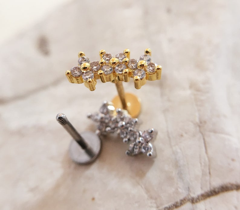 May include: Two gold and silver body jewelry pieces with clear crystal accents. The gold piece is a flower-shaped stud with a post and a clear crystal center. The silver piece is a flower-shaped stud with a post and a clear crystal center.
