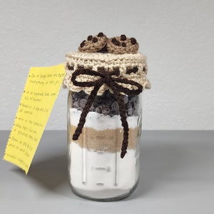 May include: A glass jar filled with cookie ingredients, topped with a crocheted lid and two crocheted chocolate chip cookies. A yellow note with instructions is visible to the left of the jar.