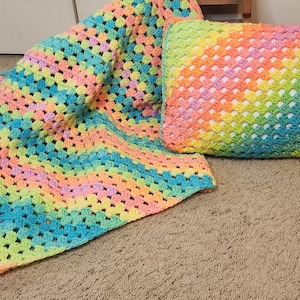 Op de afbeelding: Een regenboog gehaakte deken en kussenset. De deken is driehoekig met een kleurverloop van lichtblauw, groen, geel, roze en oranje. Het kussen is rechthoekig en heeft een vergelijkbaar kleurverloop.