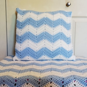 May include: A crocheted pillow and blanket set. The pillow and blanket feature a chevron pattern in light blue and white. The blanket is draped over a surface, with the pillow resting on top.