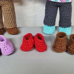 Könnte beinhalten: Sechs Paare gehäkelter Puppenschuhe in verschiedenen Farben, darunter Lila, Rot, Braun und Blau. Die Schuhe sind in drei Reihen auf einer grauen Oberfläche angeordnet.
