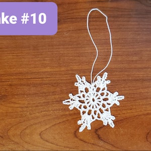 May include: White crocheted snowflake ornament with a loop for hanging. The snowflake is on a brown wooden surface. Snowflake #10.