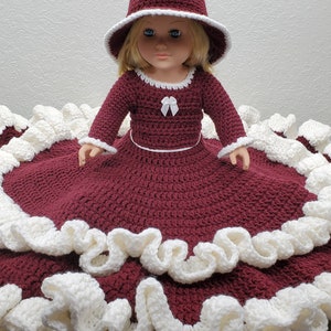 May include: A doll wearing a burgundy crocheted dress with white crocheted trim and a white crocheted hat.