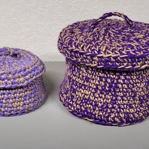 Puede incluir: Dos cestas de crochet con tapas, una más grande que la otra. Ambas cestas son de color morado y beige.