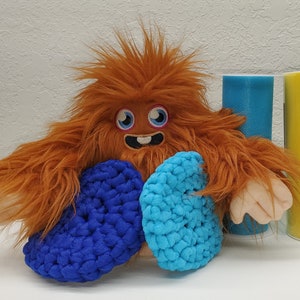 May include: A brown furry monster with blue and turquoise crocheted circles around its neck. The monster has a friendly face with big blue eyes and a toothy grin.