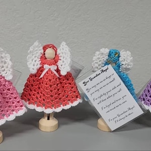 May include: Four crocheted angel figurines with white wings and dresses in pink, coral, blue, and purple. Each angel is standing on a wooden base. A small card with the text "Your Guardian Angel" is in front of one of the angels.