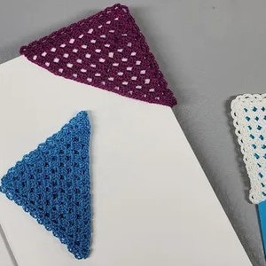 Può includere: Tre segnalibri a forma di triangolo all'uncinetto in diversi colori: viola, blu e bianco. I segnalibri sono posizionati su una superficie bianca.