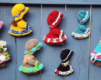Peut inclure: Sept poupées au crochet avec des robes et des chapeaux de différentes couleurs accrochés à une clôture en bois bleue. Les poupées sont toutes tournées dos à la caméra. Les poupées sont faites avec des fils de différentes couleurs, dont le rose, le rouge, le jaune, le bleu et le marron.