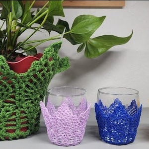 May include: Four crocheted glass covers in different colors: purple, blue, yellow, and green. The covers are placed on top of clear glass cups.