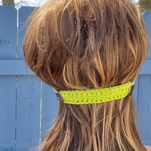 May include: A lime green crocheted mask extender with brown buttons. The extender is worn by a person with long brown hair.