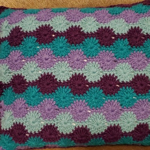 Puede incluir: Una almohada de crochet con un patrón morado, turquesa y azul claro. La almohada está hecha con un punto texturizado que crea un diseño elevado con forma de flor.