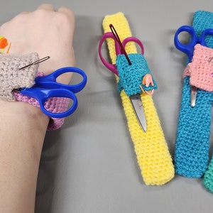 Può includere: Quattro polsini all'uncinetto per contenere gli accessori per la cucito. Un polsino è rosa e ha una piccola tasca beige con aghi e un paio di forbici. Un altro polsino è giallo e ha un paio di forbici. Il terzo polsino è blu e ha un paio di forbici e un ago. Il quarto polsino è verde e ha un paio di forbici e un ago.