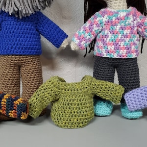 Może przedstawiać: Pięć swetrów wykonanych na szydełku w różnych kolorach i wzorach. Swetry leżą na szarej powierzchni.