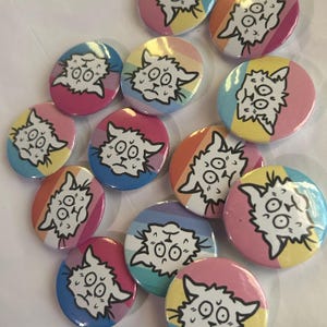 May include: A selection of circular badges displaying a cartoon cat with a concerned expression. The badges feature a white cat design with black outlines, set against a backdrop of pink, blue, yellow, and orange stripes.