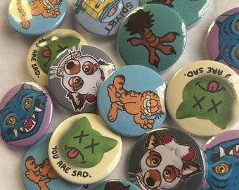 Può includere: Una collezione di spille rotonde e colorate con vari personaggi e disegni di cartoni animati. Le spille presentano Garfield, SpongeBob e altre illustrazioni stravaganti. Alcune spille hanno il testo "YOU ARE SAD." e "SDIYBT."