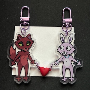 Peut inclure: Deux breloques en acrylique représentant un renard marron et un lapin violet clair, chacune dotée d'un fermoir mousqueton en métal violet. Les deux personnages sont reliés par un petit cœur rouge, formant une paire d'accessoires décoratifs.