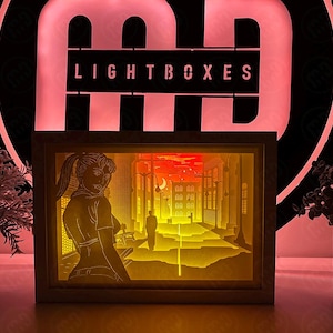Puede incluir: Una caja de luz de papel 3D con una mujer en una escena de ciudad. La mujer lleva una camisa blanca y tiene el pelo recogido hacia atrás. La escena de la ciudad está iluminada con luces amarillas. La caja de luz está hecha de madera y tiene un marco blanco.