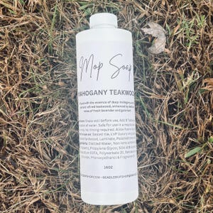 May include: A white bottle of "Mop Soap" with a label that reads "Mahogany Teakwood." The bottle is sitting on a bed of dry grass. The label includes instructions and ingredients. The bottle contains 16 oz of soap.