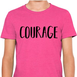 以下が含まれることがあります： ピンクのTシャツに黒い文字で「COURAGE」とプリントされています