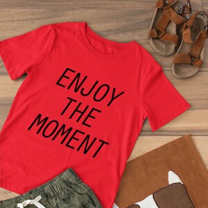 以下が含まれることがあります： 黒文字で「Enjoy the Moment」とプリントされた赤い半袖Tシャツ。緑色の迷彩柄のショートパンツと茶色の犬の形をしたクッションと組み合わせています。