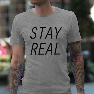 以下が含まれることがあります： 黒い文字で「STAY REAL」とプリントされたグレーのTシャツです。