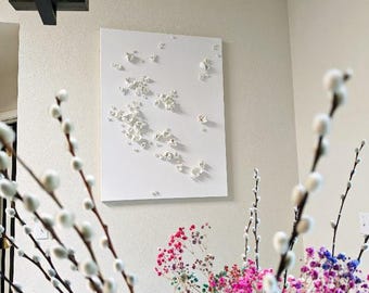 May include: A rectangular white wall art piece featuring a textured, raised relief design of small, scattered white floral or organic shapes. The artwork is mounted on a plain wall, positioned below a dark brown wooden light fixture.