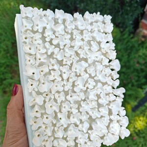 May include: A white, textured art piece featuring a repeating pattern of abstract, flower-like shapes. The artwork is held in a hand, showcasing the three-dimensional relief of the design. The background is blurred, with green foliage.