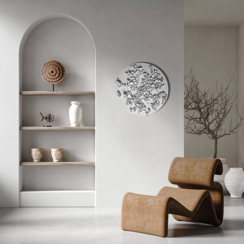 May include: A white, round wall sculpture with a textured, organic design. A brown leather armchair sits in front of the sculpture. A wooden shelf with decorative items is in the background.
