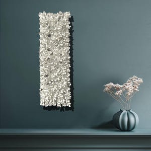 May include: A white ceramic wall sculpture with a textured, organic design. The sculpture is mounted on a teal blue wall, and a small vase with dried flowers sits on a shelf below.