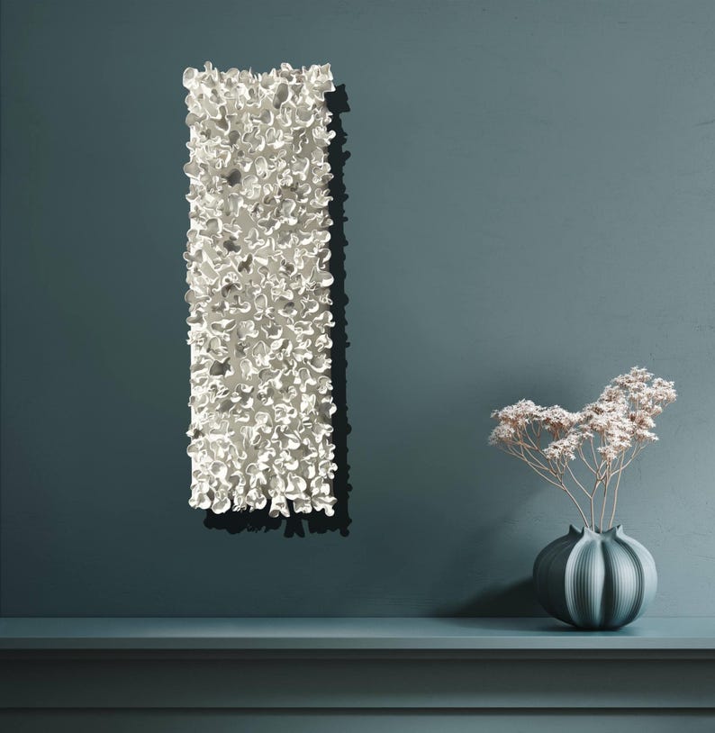 May include: A white ceramic wall sculpture with a textured, organic design. The sculpture is mounted on a teal blue wall, and a small vase with dried flowers sits on a shelf below.
