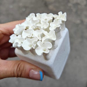May include: A white ceramic sculpture of a cluster of small, ruffled flowers arranged in a cube shape.