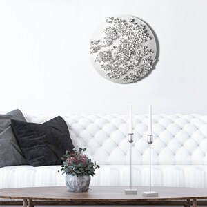 May include: A white, round wall sculpture with a textured, organic design. The sculpture is hanging on a white wall above a white sofa with black pillows. There are two silver candle holders with white candles on a wooden coffee table in front of the sofa.