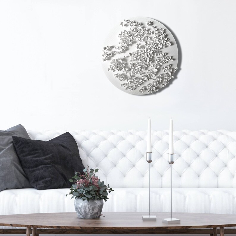 May include: A white, round wall sculpture with a textured, organic design. The sculpture is hanging on a white wall above a white sofa with black pillows. There are two silver candle holders with white candles on a wooden coffee table in front of the sofa.