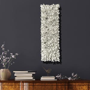May include: White ceramic wall sculpture with a textured, organic design. The sculpture is hanging on a dark gray wall above a wooden dresser with books and decorative items.