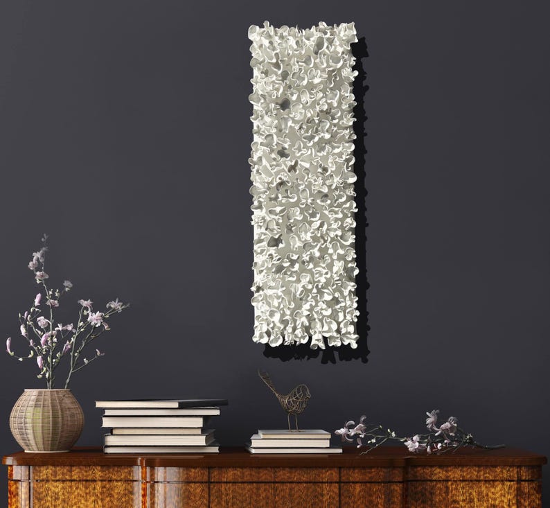 May include: White ceramic wall sculpture with a textured, organic design. The sculpture is hanging on a dark gray wall above a wooden dresser with books and decorative items.