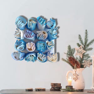May include: A wall-mounted art piece featuring a grid of blue and white abstract forms. The forms have a layered, textured appearance, with a mix of light and dark blue hues. The artwork is displayed on a white background.