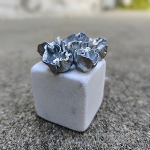 May include: A small cluster of silver-colored metal flowers sits atop a white cube. The flowers have a delicate, ruffled texture.