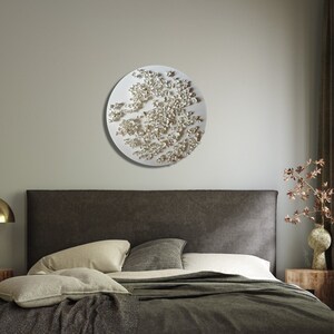 May include: A white circular wall sculpture with a textured, raised design resembling a cluster of small white flowers. The sculpture is hanging on a light gray wall above a brown headboard.