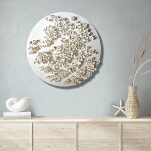 May include: White ceramic wall sculpture with a circular shape and a textured surface. The sculpture is made up of many small, organic shapes that resemble flowers or leaves. It is hanging on a light blue wall above a wooden shelf.