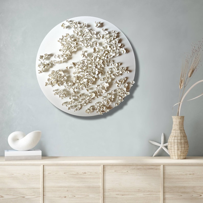 May include: White ceramic wall sculpture with a circular shape and a textured surface. The sculpture is made up of many small, organic shapes that resemble flowers or leaves. It is hanging on a light blue wall above a wooden shelf.